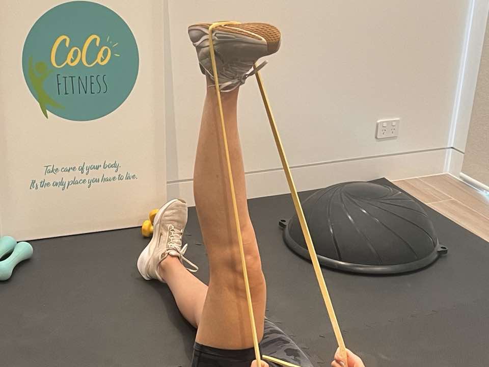 A resistance-band leg exercise on a floor mat in a home room, with a CoCo Fitness banner reading &lsquo;Take care of your body. It&rsquo;s the only place you have to live&rsquo;, plus dumbbells and a balance trainer beside the mat.