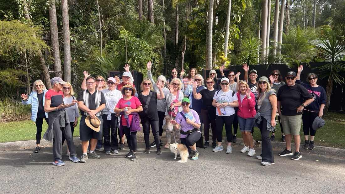 About twenty-five women of the Noosa Walk and Talk group gathered on a quiet road before a walk, many waving at the camera, rainforest behind them and a small dog at the front.