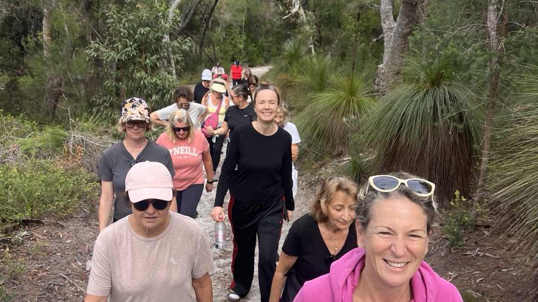 The Noosa Walk and Talk group walking a sandy track through coastal bushland, a line of women of different ages stretching away up the trail, Corinna nearest the camera in a pink hoodie.